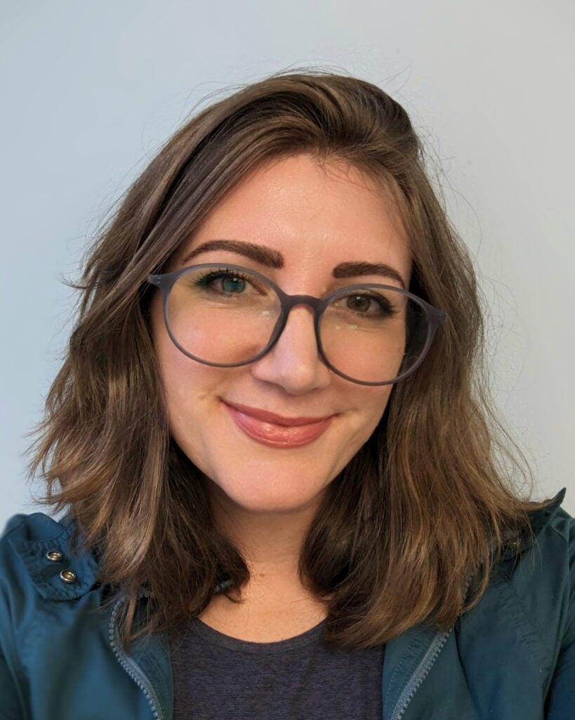 A woman with shoulder-length brown hair, wearing large round glasses and a blue jacket, smiles at the camera against a light background.