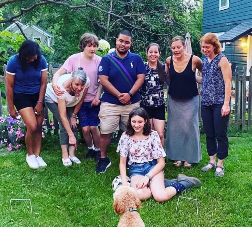 A group of eight people pose outside on grass, some standing and some crouching; a dog sits facing a seated girl in the center. There are trees and a house in the background.