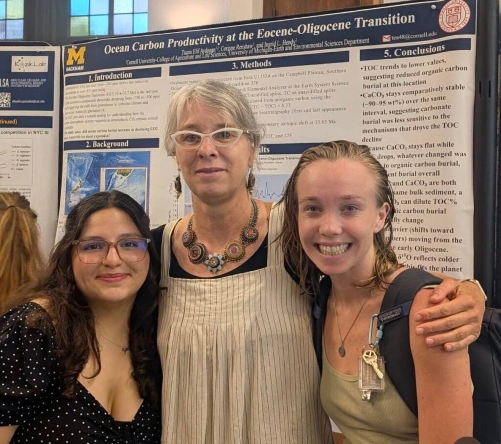 Three people stand smiling in front of a scientific research poster. Two younger women stand on either side of an older woman with short gray hair and glasses. The poster discusses ocean carbon productivity.