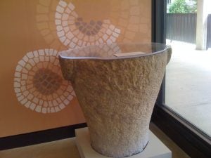 A large, ancient stone basin is displayed on a pedestal indoors. Behind it, a wall features a mosaic pattern with circular designs in beige and brown tones.