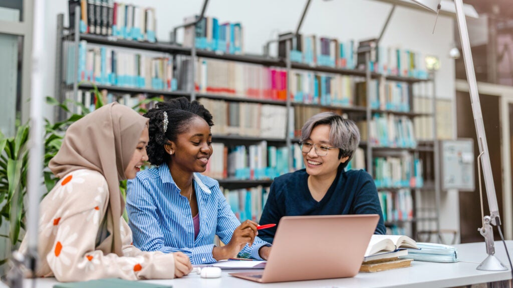 Three students in the library working together on a laptop.