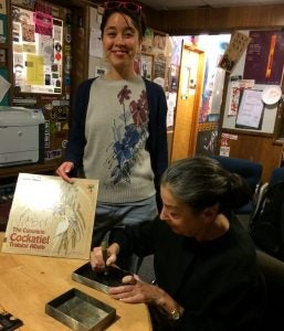 the artist signs her photobook and the engineer, Liz, smiles while holding a record album The Cocktiel