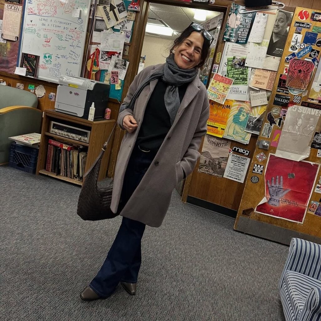 A smiling woman, possibly Natalie Bakopoulos, stands in an office with walls covered in colorful posters and notes. She wears a gray coat, scarf, dark pants, and carries a woven bag, with one hand in her pocket.