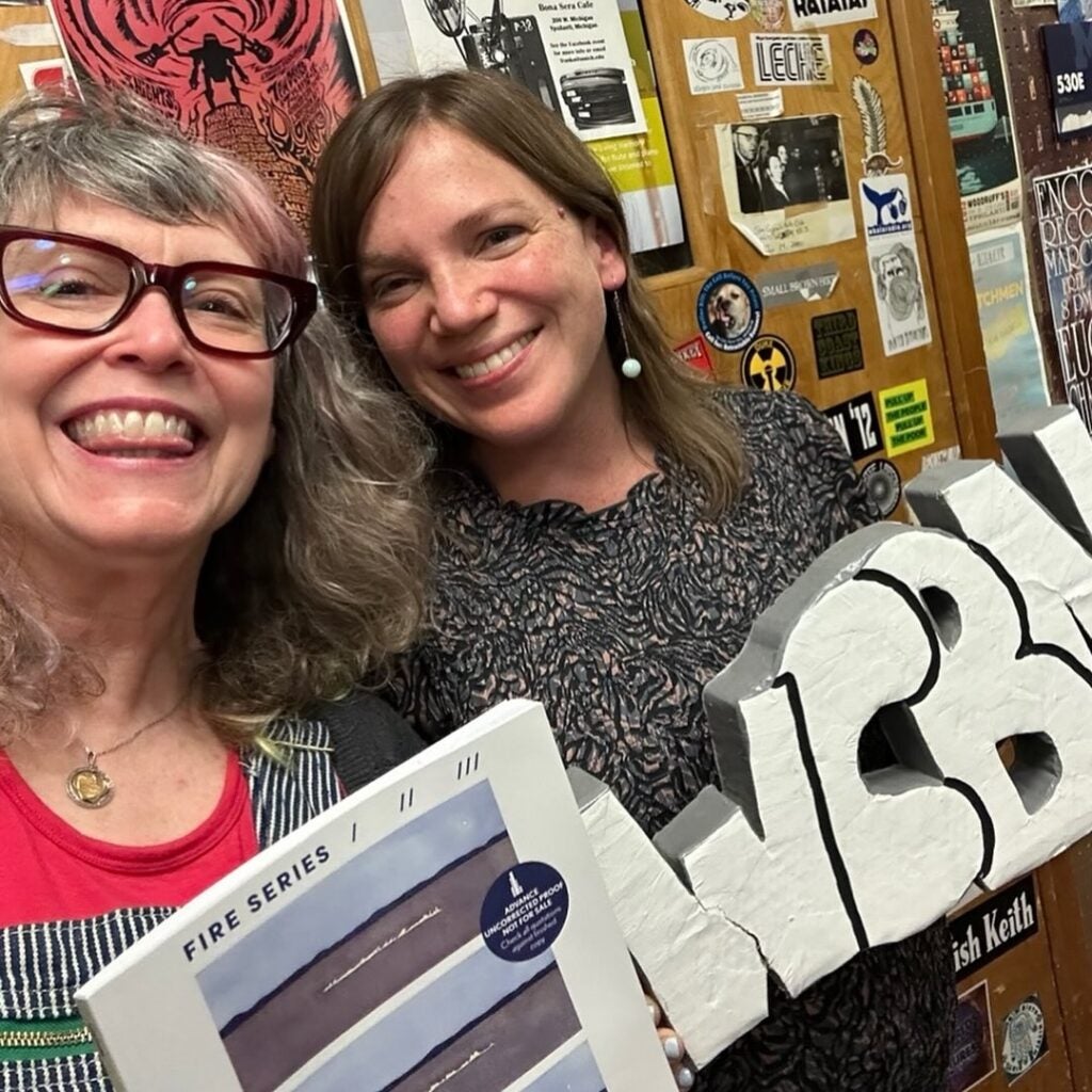 Two smiling women pose indoors, one holding a book and the other holding a large cutout of the letters WCBN. Behind them is a bulletin board covered with posters and stickers, hinting at an event featuring Kelly Hoffer.