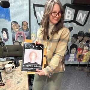 A woman wearing glasses and a plaid jacket holds up Jennifer Sperry Steinorth’s book, Boys Behind Glass, in a radio studio adorned with a mural of famous musicians and a WNBN sign in the background.