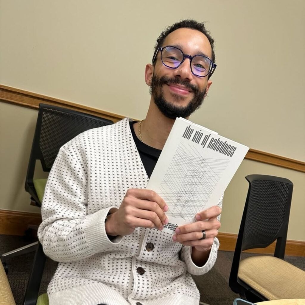 A smiling person wearing glasses and a cream-colored sweater sits in an office chair, holding up a book titled The King of Confidence by Chris Crowder.