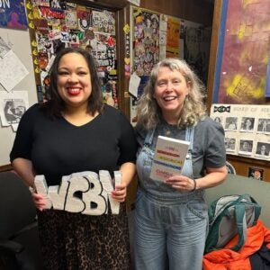 Two women smile in a radio studio. One holds large “WCBN” letters, while the other—Danielle Evans—holds a book titled The Somewhat Historical Irreverence of Danielle. The background is filled with posters, stickers, and photos on the walls.