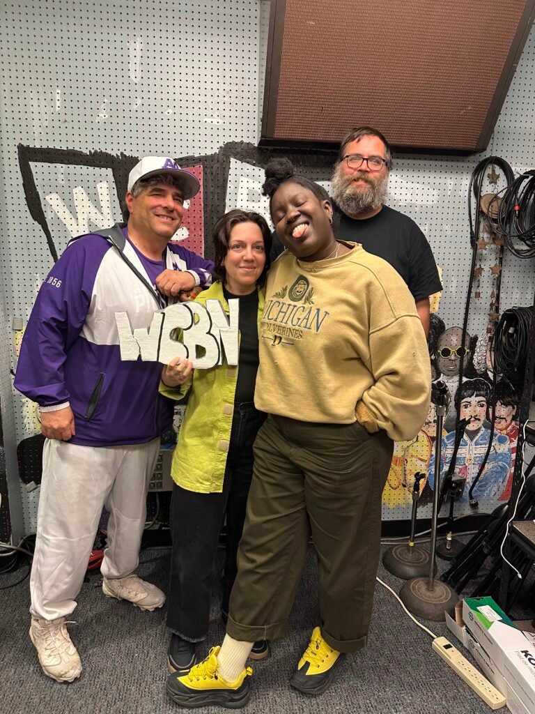 Shira Erlichman, Angel Nafis, and Jeff Kass smile and pose with another guest in a radio station studio. One person holds up a WCBN sign amid equipment, cables, and colorful wall art in the background.