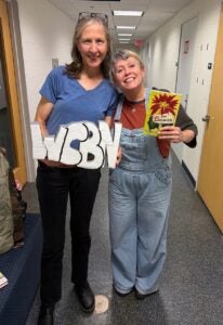 Two women smiling in a hallway, one holding a foam cutout of the letters WBN and the other holding a book titled The Spirits by Bonnie Jo Stufflebeam, which is also written by Bonnie Jo Williams.