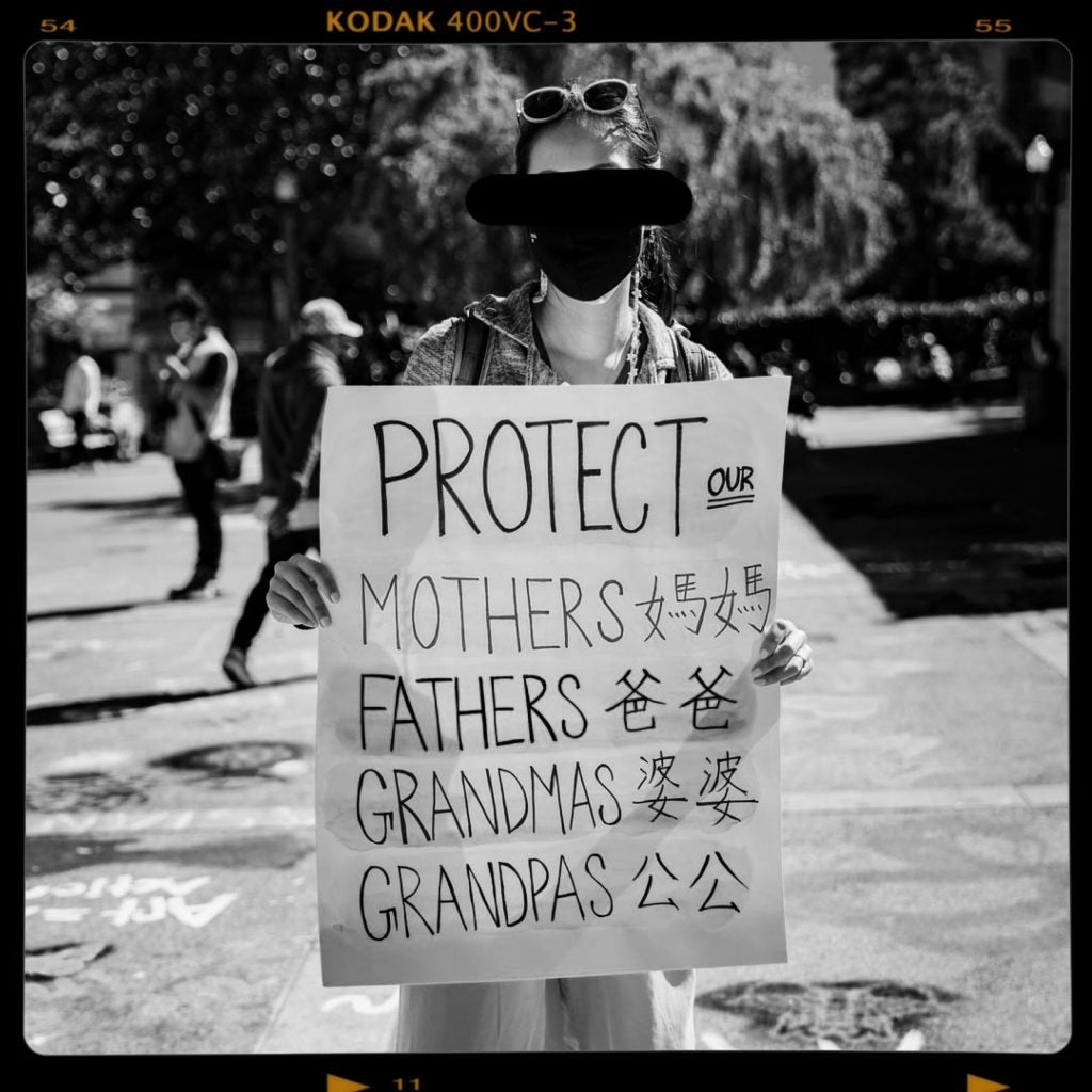 A black and white photograph if a woman at a protest who is facing the camera and holding up a sign that says "protect our mothers, fathers, grandmas, grandpas." The poster includes translations for the words mother, father, grandma, and grandpa in Chinese next to the English words. A black line has been drawn through the woman's eyes in the photograph to signify her anonymity.