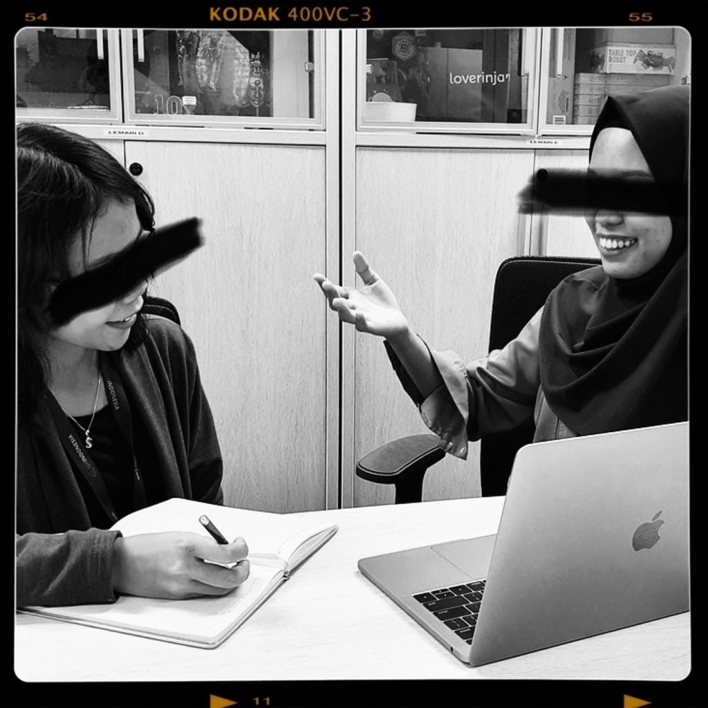 A black and white photograph of two women sitting at a conference table and facing each other. The woman on the left is taking notes in a notebook and looking up at the other smiling. There is a laptop in front of the other woman on the right which positioned so they both can see the screen. The woman on the right has her right arm raised as if making a point as she smiles at the woman on the left. A black line has been drawn through both women's eyes in the photograph to signify their anonymity.