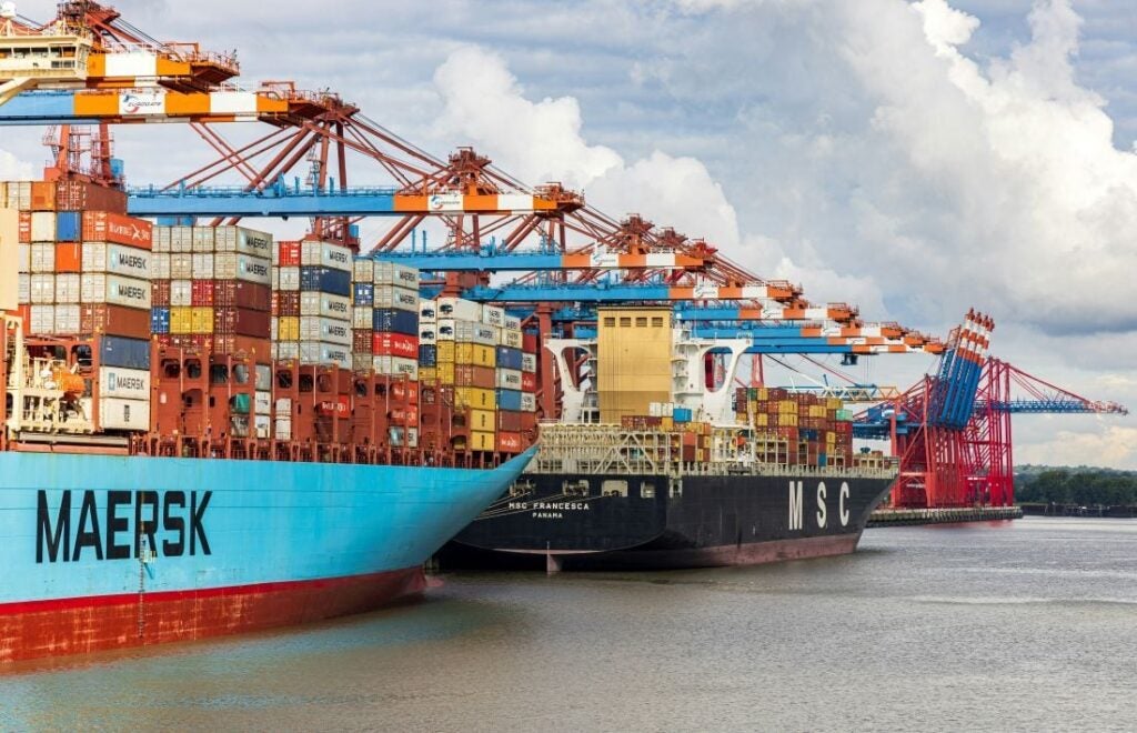 Two large container ships docked at a busy port with colorful cranes and stacked shipping containers along the waterfront under a partly cloudy sky.