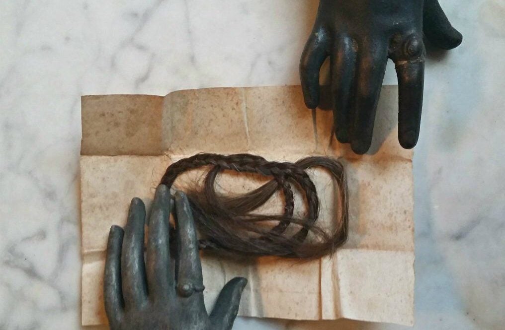 two ceramic hands, one black, one grey, holding open a piece of paper with hair in the middle