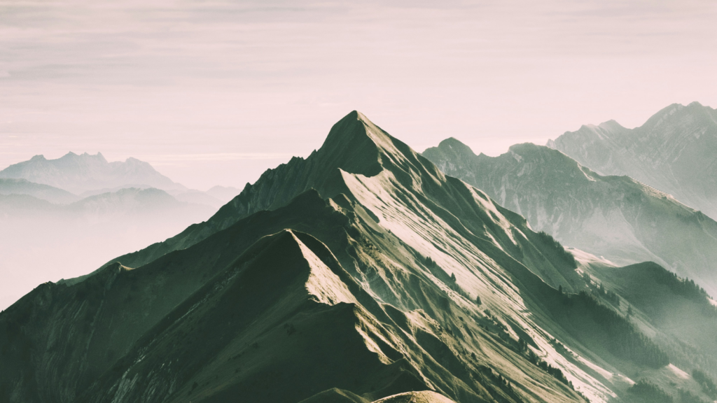 A rugged mountain ridge with sharp peaks stretches into the distance, bathed in soft light and surrounded by mist, with layers of mountains and empty summits visible in the background.