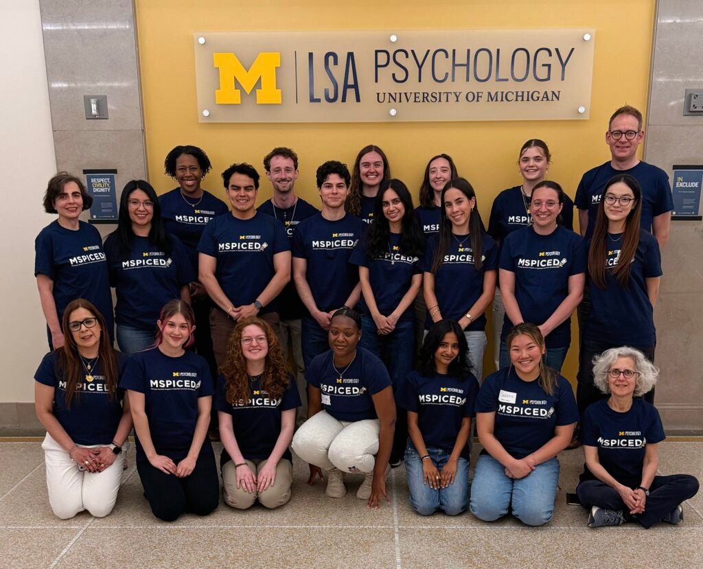 Group of 20 people wearing matching navy blue “MSPICED” shirts pose and smile in front of a yellow and white University of Michigan LSA Psychology sign indoors.