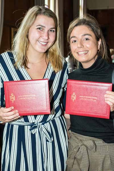 Two recent inductees wearing dresses smile at the camera while holding up their red Alpha of Michigan Chapter certificate folders.