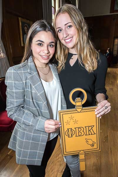 Two long-haired inductees, one in a plaid bazer and white blouse and the other in a black blouse, hold up a gold Phi Beta Kappa key and smile at the camera.
