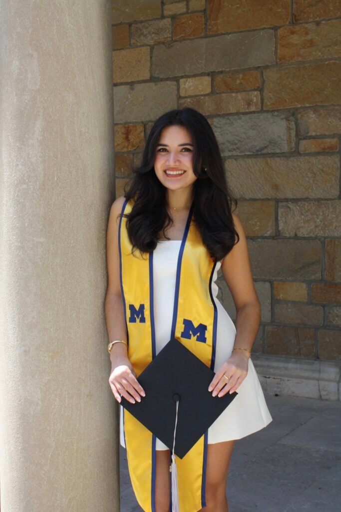 A young woman in a white dress and yellow graduation stole stands by a stone pillar, smiling and holding a graduation cap with a tassel. The stole features blue M letters, and a stone wall is in the background.