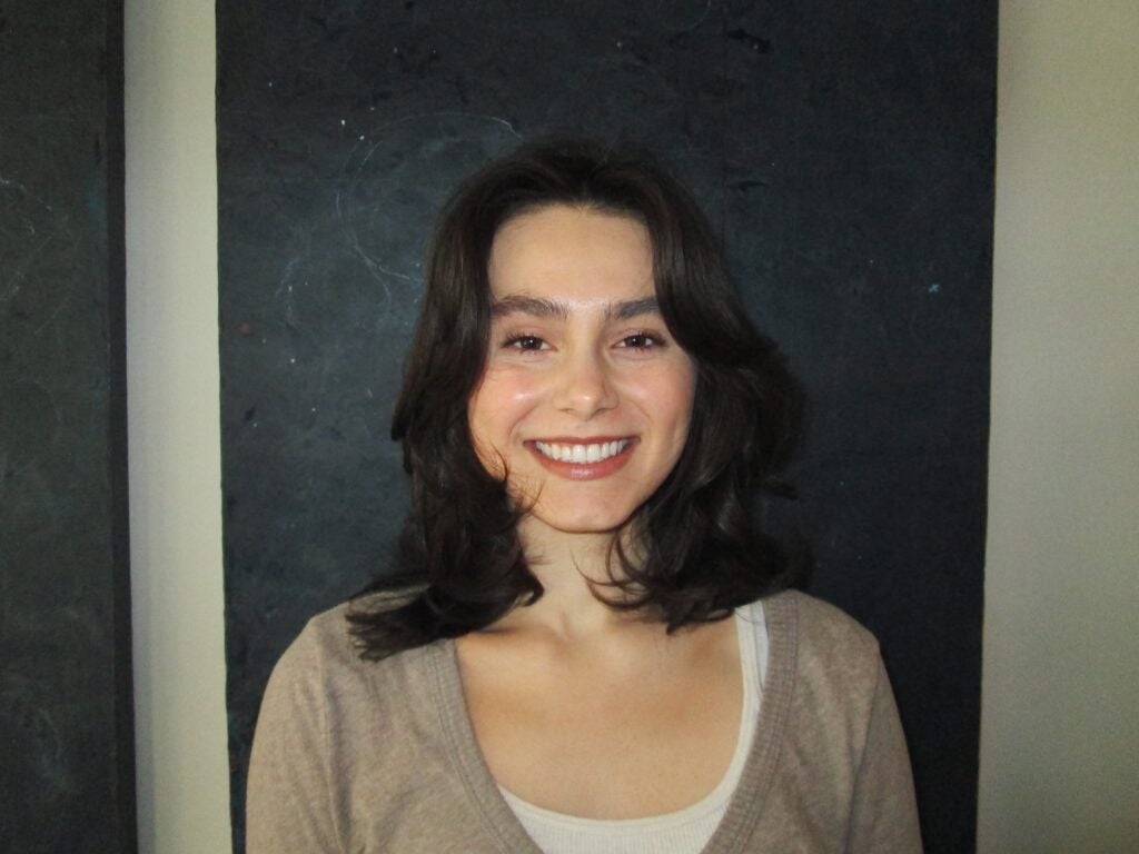 A young woman with medium-length dark hair smiles at the camera. She is wearing a light tan sweater over a white top, and is standing in front of a dark, textured background.