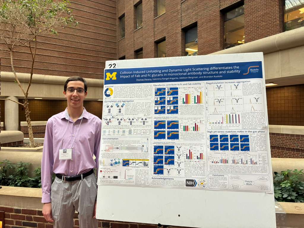 Thomas, wearing glasses and a name badge, stands beside his scientific research poster at an indoor URS event. The poster features graphs, charts, and text, with a University of Michigan logo at the top left corner.