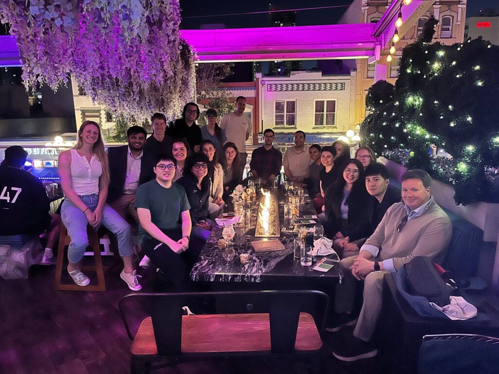 A group of people pose and smile around a long dinner table at an outdoor rooftop restaurant at night, with purple lighting, hanging plants above, and city buildings in the background.