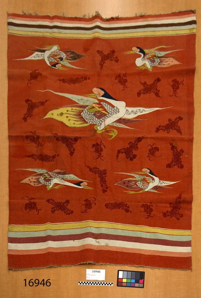 A reddish-orange textile featuring colorful, stylized birds in flight and floral patterns. The bottom has multicolored horizontal stripes. A number “16946” and a color chart are visible at the bottom.