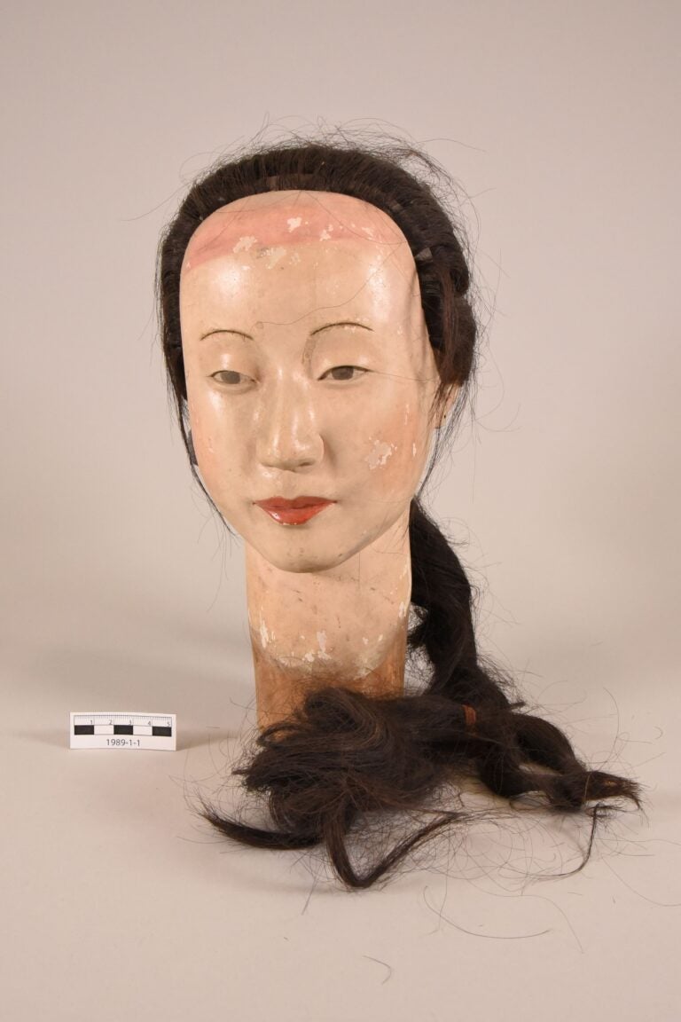 A mannequin head with painted facial features and real dark hair in a braid. The mannequin shows signs of wear, with paint chipping on the face. A small scale for measurement is placed beside it.
