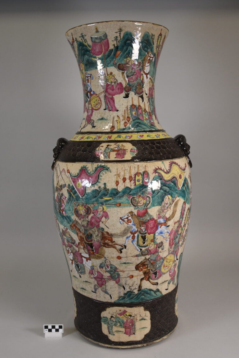 Tall ceramic vase with a flared rim and intricate, colorful painted scenes of figures in traditional attire, some holding weapons; black and white scale for size reference is placed in front.