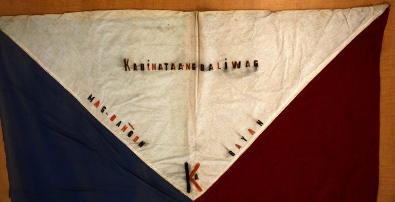 A close up image of the top of the flag, specifically the white triangle. In the middle of the triangle are the words "kabinataang baliwag." On the edges of the triangle are the words, "Mag-bangon Bayan" and a K with a small A inscribed into it.