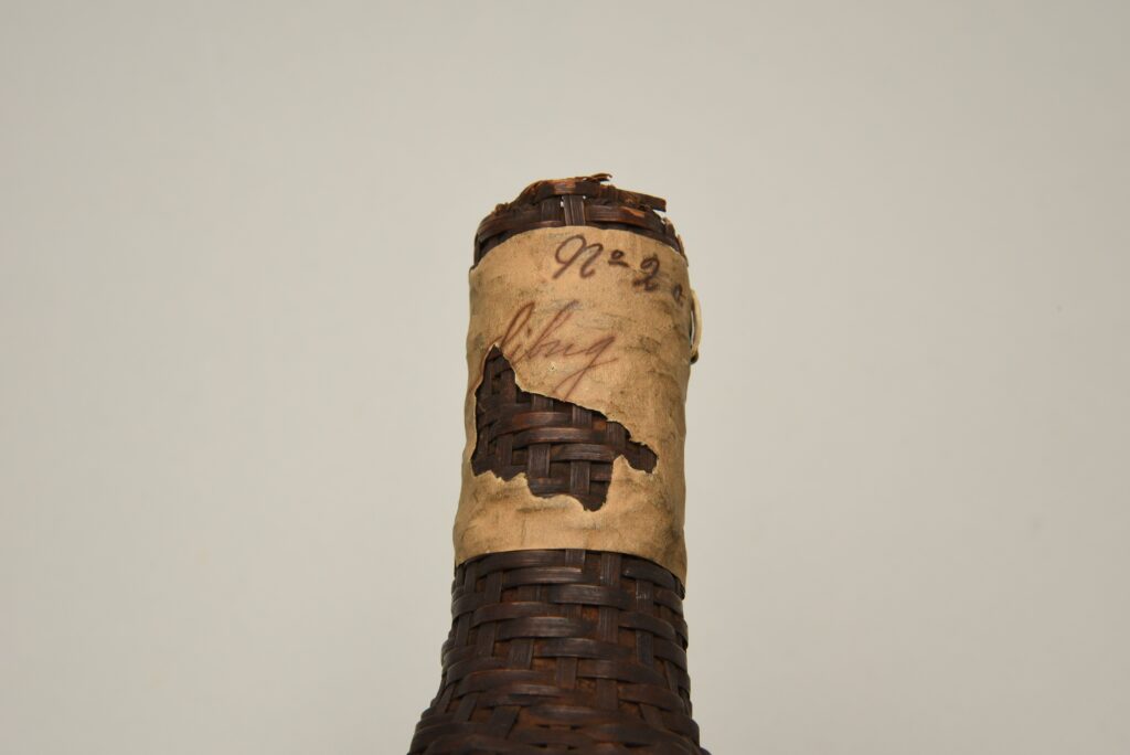 Image of the neck of basketry woven in the form a bottle, with an aged and torn label that appears to have the word "Sibug" written in cursive. Above this is written "No. 2 a" in cursive.
