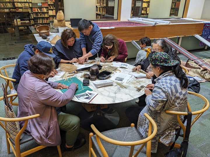 This image shows a group of people seated around a circular table in a library participating in a basket weaving activity. The participants are working with thin plant fibers or rattan strips, shaping and weaving them into small basket forms. Various tools, photographs, and weaving materials are spread across the table as reference and working materials.