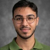 A young man with short dark hair, a beard, and glasses smiles slightly. Adel Kamel Kaddoumi is wearing a green collared shirt and stands in front of a gray, softly blurred background.
